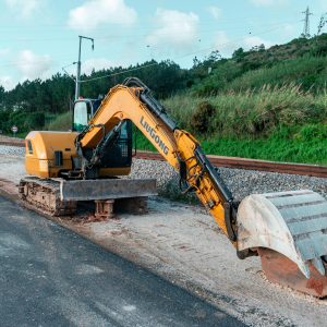 Yellow excavator parked on a construction site beside railroad tracks and greenery.